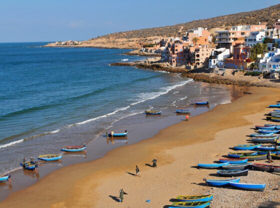 Aerial view of Taghazout, Morocco, with a village built into a hillside overlooking a beach dotted with blue and yellow fishing boats.