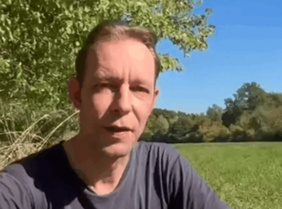 A man wearing a dark t-shirt speaking to the camera outdoors with green foliage and blue sky in the background.