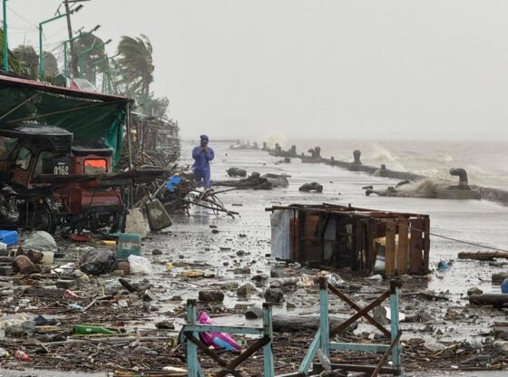 A man in a raincoat standing on a debris-strewn waterfront road with large waves crashing in the background due to Super Typhoon Ragasa.