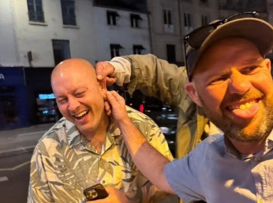 Daniel Ashley-Laws having his ear pulled by an unseen person while another man with a cap and sunglasses smiles and sticks out his tongue.