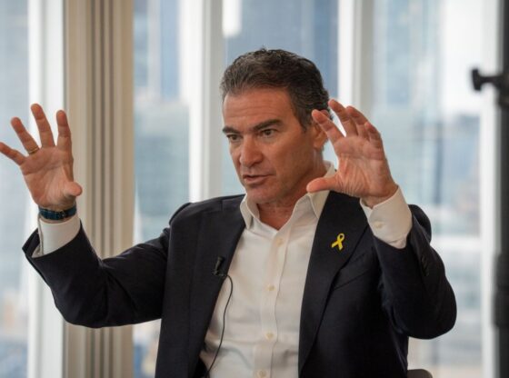 Yossi Cohen, former head of Mossad, in a suit with a yellow ribbon, gestures with both hands during an interview.
