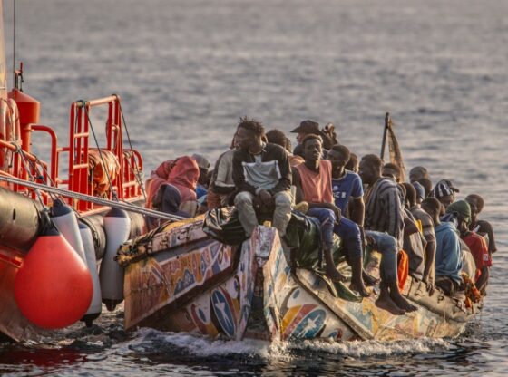 Migrants on a colorful boat are assisted by a red Spanish search and rescue vessel at sea.