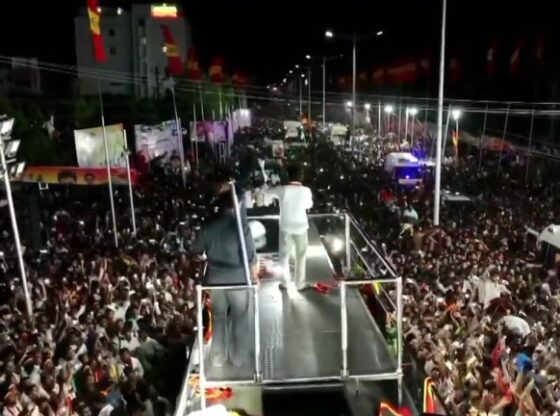 A politician addresses a large crowd from a platform on a stage at night.