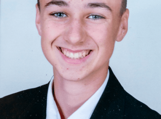 A smiling young man in a black suit and red tie with a white shirt.