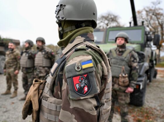 Ukrainian soldier wearing a camouflage uniform, helmet, and tactical vest, with a Ukrainian flag patch and a red and black patch with a scorpion and a white cross on the sleeve, standing next to military vehicles and other soldiers.