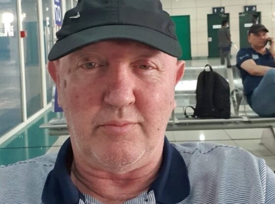 John Murphy, a British veteran, wearing a black cap and striped shirt, sitting in what appears to be an airport or public waiting area.