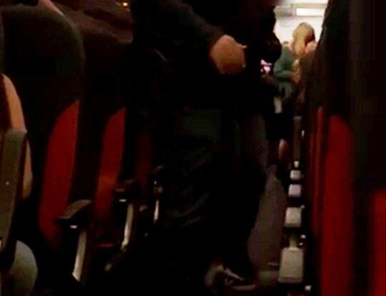 A man in a dark uniform escorts a passenger clutching a vodka bottle down an airplane aisle.