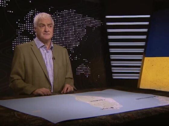 A man in a suit jacket stands in front of a news desk with a map of South America, and a backdrop displaying a world map and a cracked image of the Ukrainian and Russian flags.