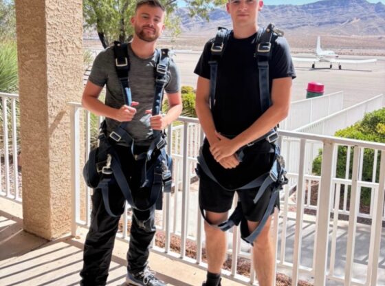 Two men in skydiving harnesses pose for a photo with a small airplane and mountains in the background.