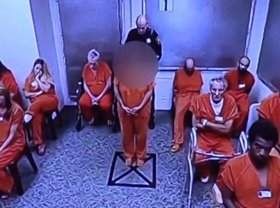 A woman in an orange jumpsuit stands facing forward while a security guard stands behind her in a room with other people in orange jumpsuits.