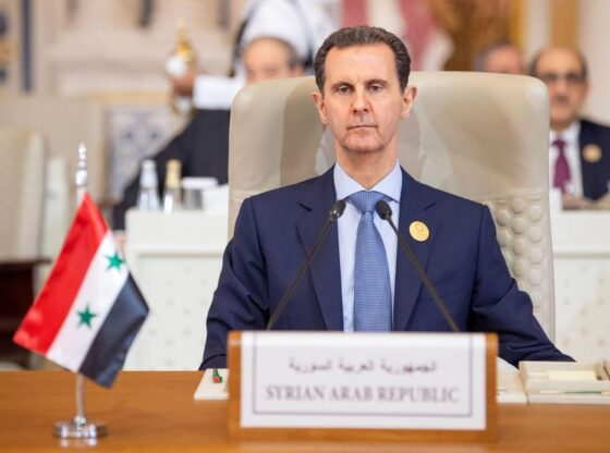 Syrian President Bashar al-Assad seated at an emergency meeting, with the Syrian flag and a nameplate reading "Syrian Arab Republic" in front of him.