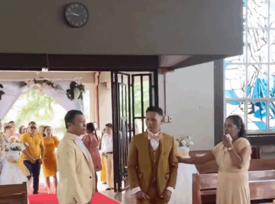 A man in a gold suit, standing on a red aisle, looks toward a woman in a long beige dress speaking into a microphone. Another man in a light suit stands to the left of the gold-suited man, looking towards a wedding procession entering through open doors.