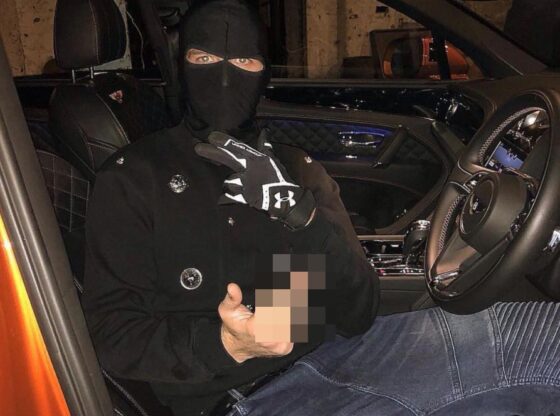 A person in a black balaclava, black jacket, and gloves, sits in the driver's seat of an orange car, holding a blurred object in one hand.