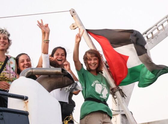 Greta Thunberg and a crew member flash victory signs from their ship, with the Palestinian flag flying.