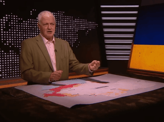 A man reporting on the news with a map of Ukraine on a table in front of him, and an image of the Ukrainian flag behind him.