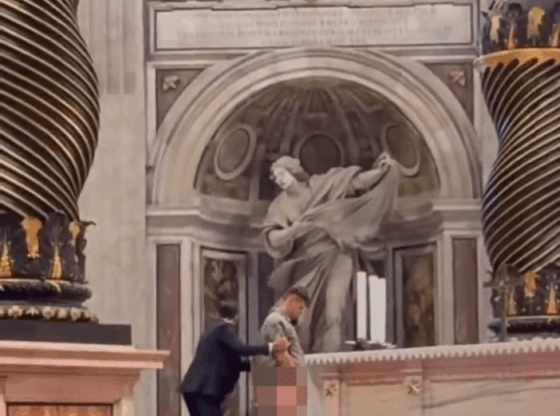 A man in a light grey outfit with his pants down is being restrained by another man in a dark suit in front of an altar, with his privates pixelated.