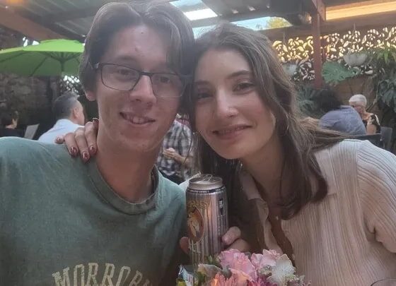 A couple poses at an outdoor restaurant, the woman holding a can and a bouquet of flowers.
